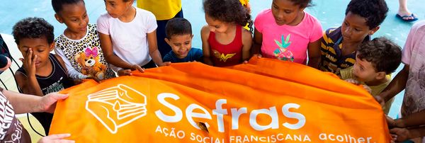 A imagem mostra um grupo de crianças reunidas ao redor de um grande tecido laranja com o logotipo Sefras – Ação Social Franciscana estampado. Elas estão posicionadas lado a lado, segurando a parte superior do tecido. O ambiente parece ser uma área aberta ou quadra, com piso azul ao fundo. As crianças vestem roupas coloridas e diversas, algumas com estampas infantis e outras com personagens. A cena transmite um clima de participação coletiva e atividade comunitária.
